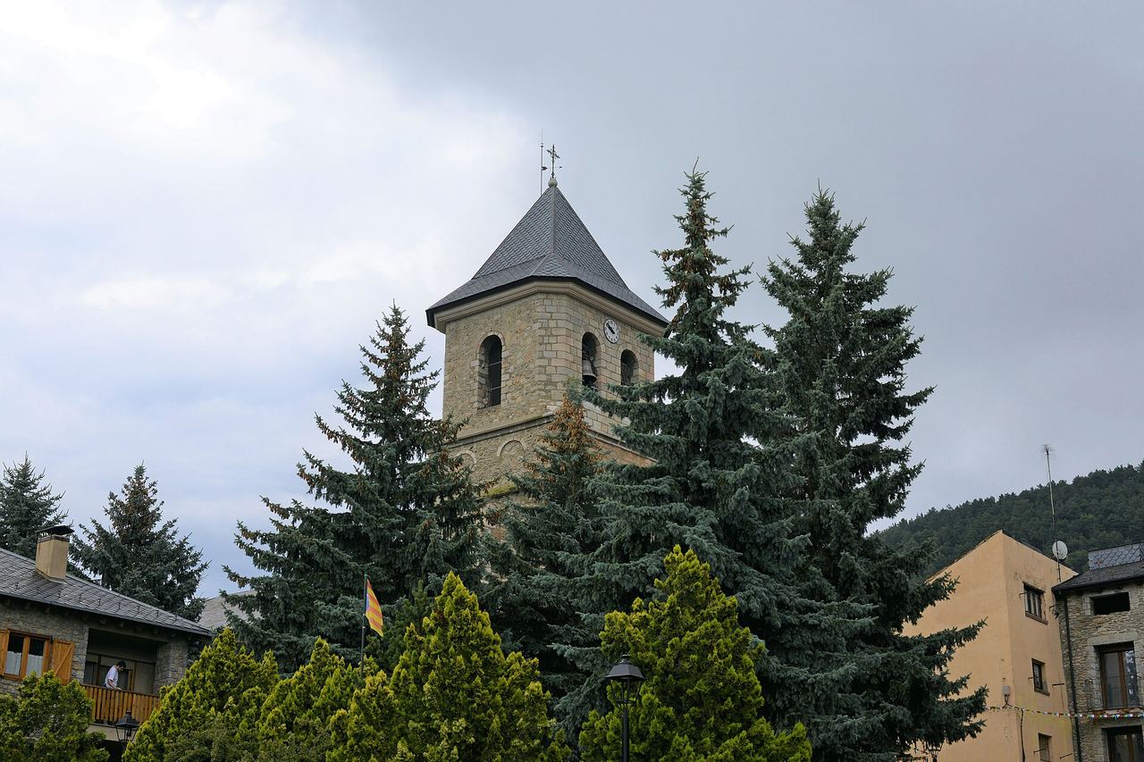 Campanar de pedra de Sant Pere d'Alp, amb rellotge i teulada de pissarra, entre coníferes altes sota un cel ennuvolat
