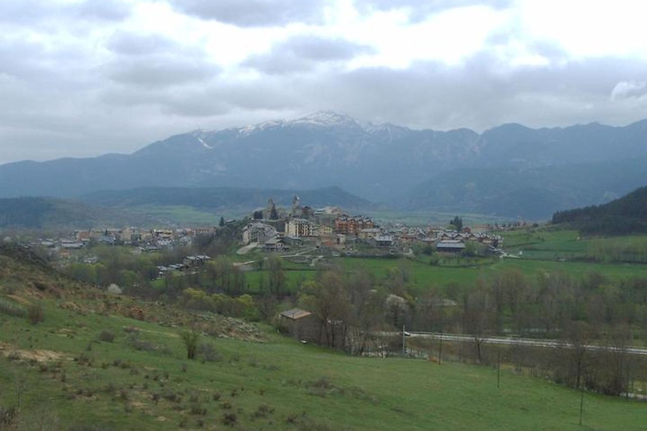 Bellver de Cerdanya dalt d'un turó, entre prats verds, amb muntanyes de cims nevats i núvols al fons