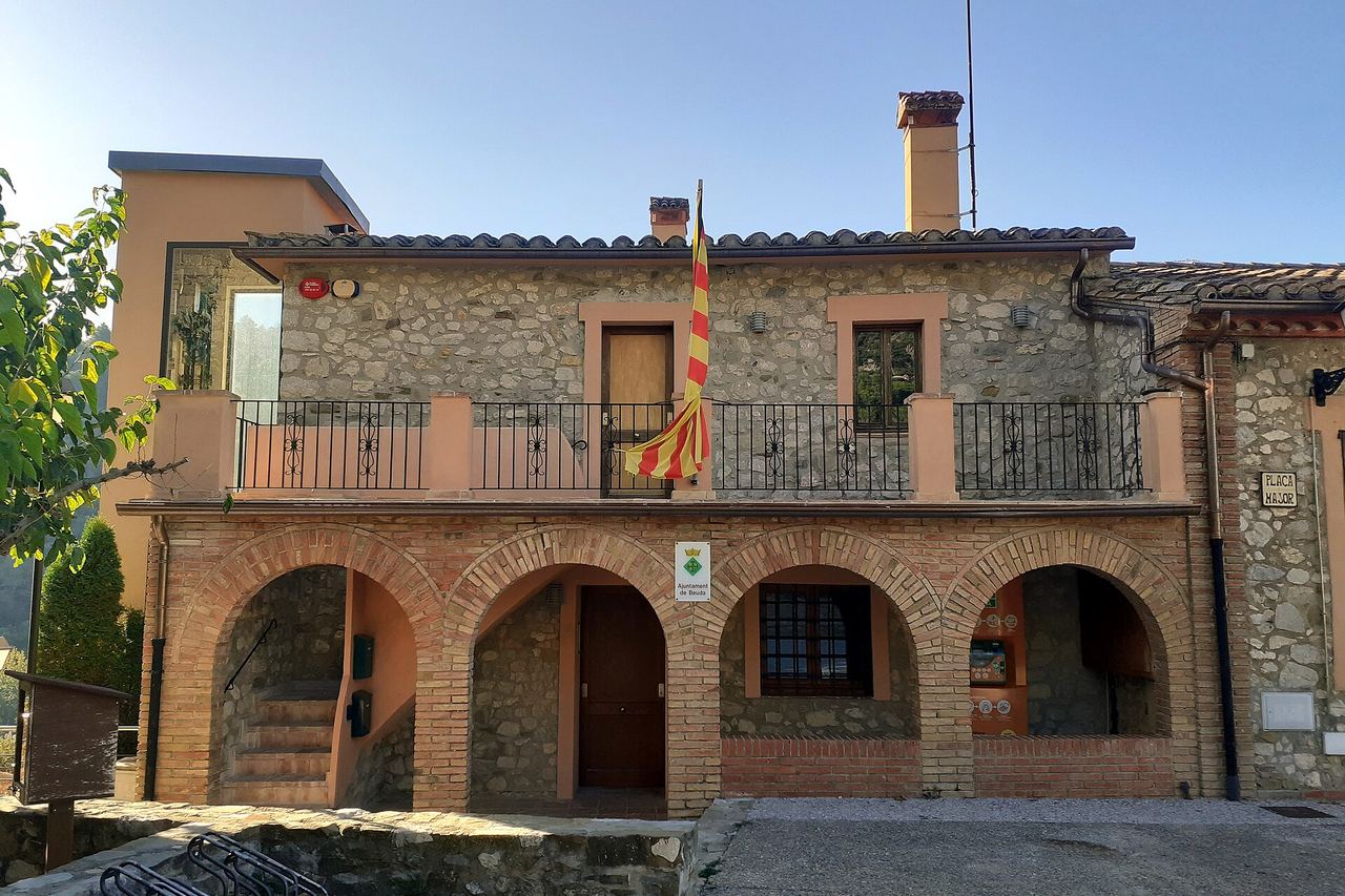 La casa de la vila de Beuda, de pedra i maó, amb quatre arcs als baixos, balcó de ferro i la senyera, a la plaça Major