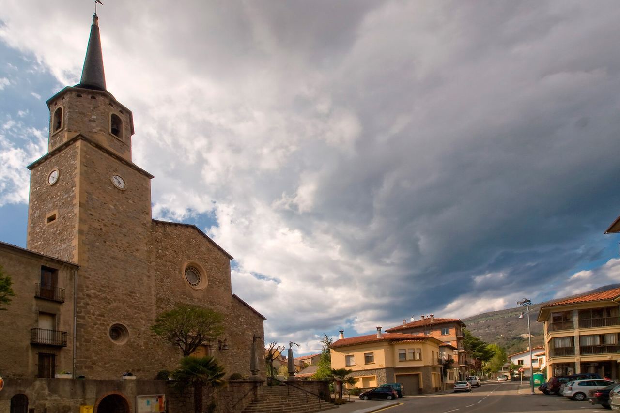 Església de Campdevànol amb el campanar de pedra, rellotges i agulla, al costat d'un carrer amb cases i cotxes