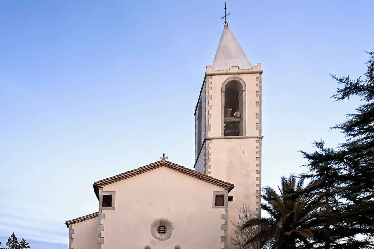 L'església de Sant Quirze de Campllong, de façana clara, amb el campanar acabat en punxa i una palmera al costat