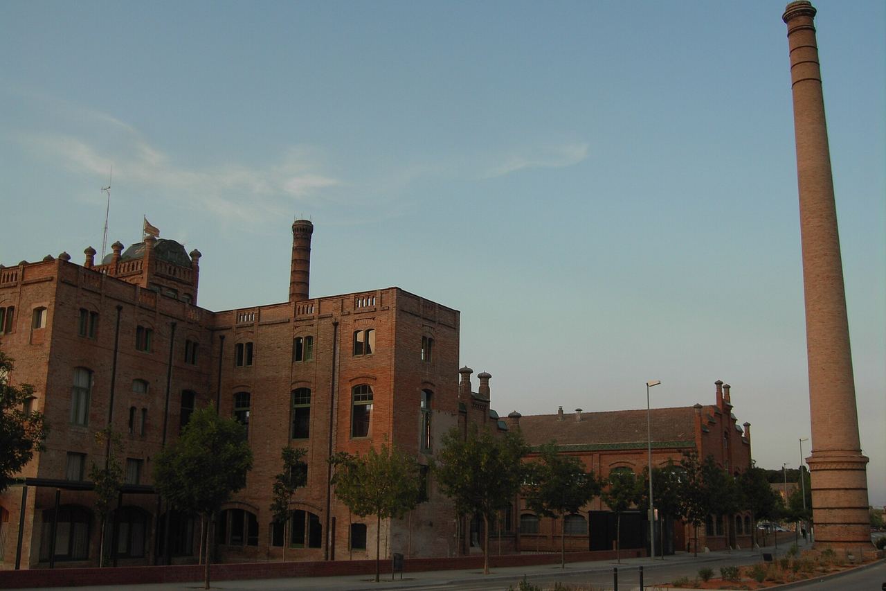 La Fàbrica de Celrà, un gran edifici de maó amb finestres d'arc, i una xemeneia de maó molt alta a la dreta