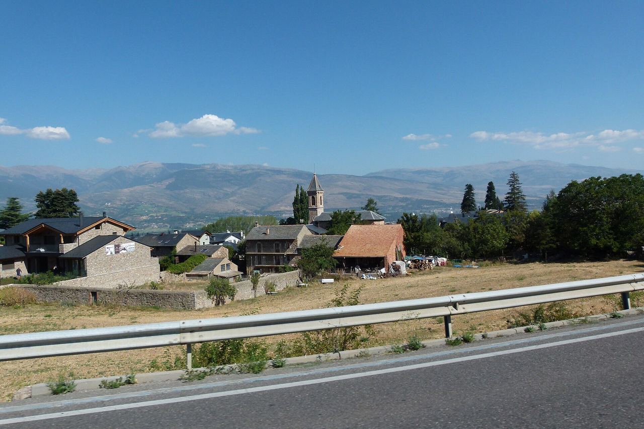 Das des de la carretera: un camp sec, l'església amb campanar punxegut, cases de pedra i muntanyes sota un cel blau