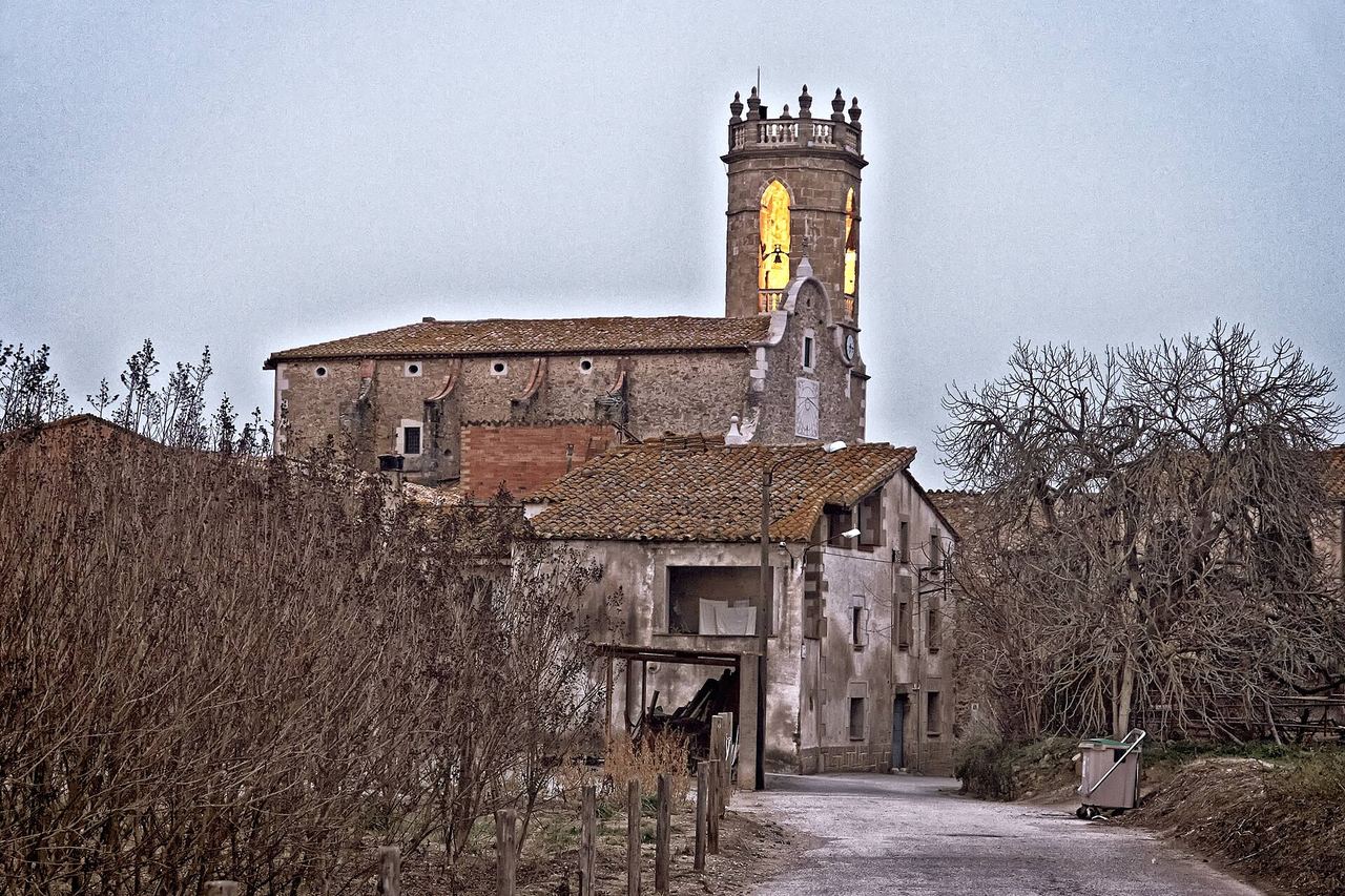 Un camí de Flaçà amb una casa antiga, arbres sense fulles i, al darrere, l'església amb el campanar coronat de pinacles