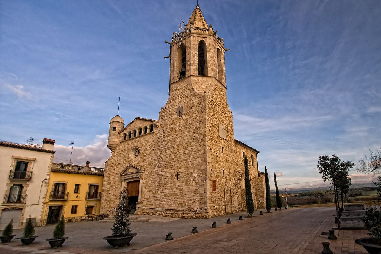 L'església de Sant Cugat, a Fornells de la Selva, amb un campanar alt de pedra, xiprers i una plaça amb pilones al davant