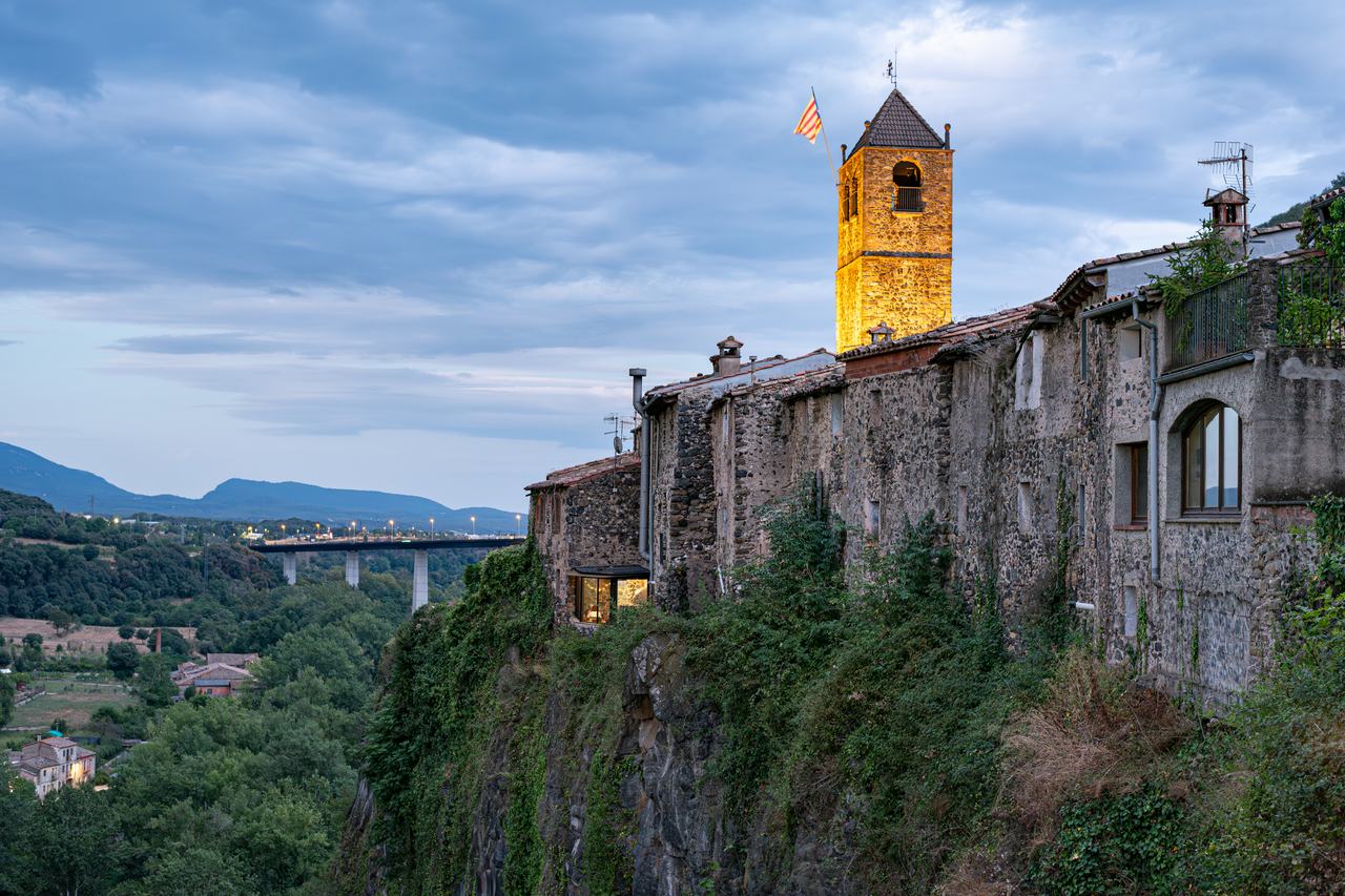 El poble de Castellfollit de la Roca dalt del cingle de basalt, amb el campanar il·luminat al capvespre