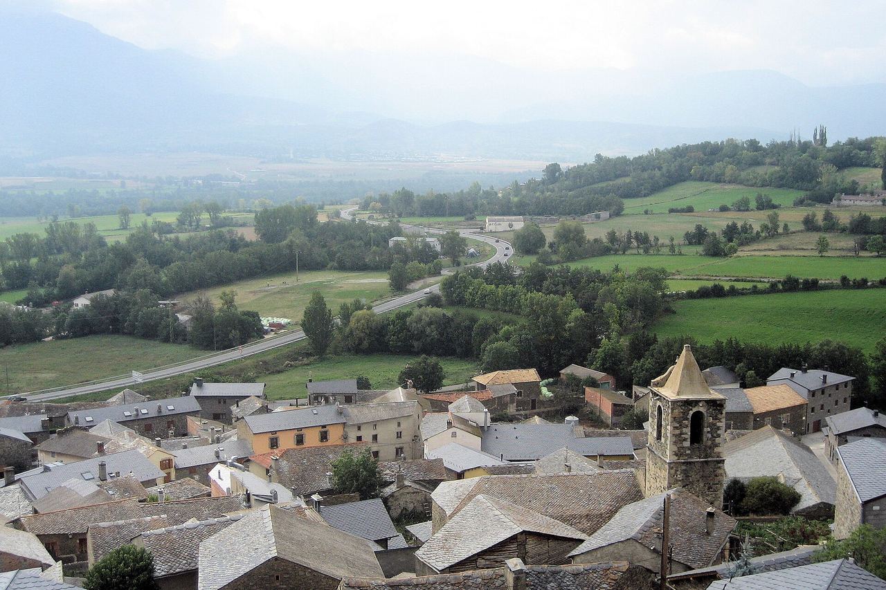 Teulades de pissarra i el campanar de pedra de Ger, amb la vall de la Cerdanya al fons: prats, arbres i una carretera
