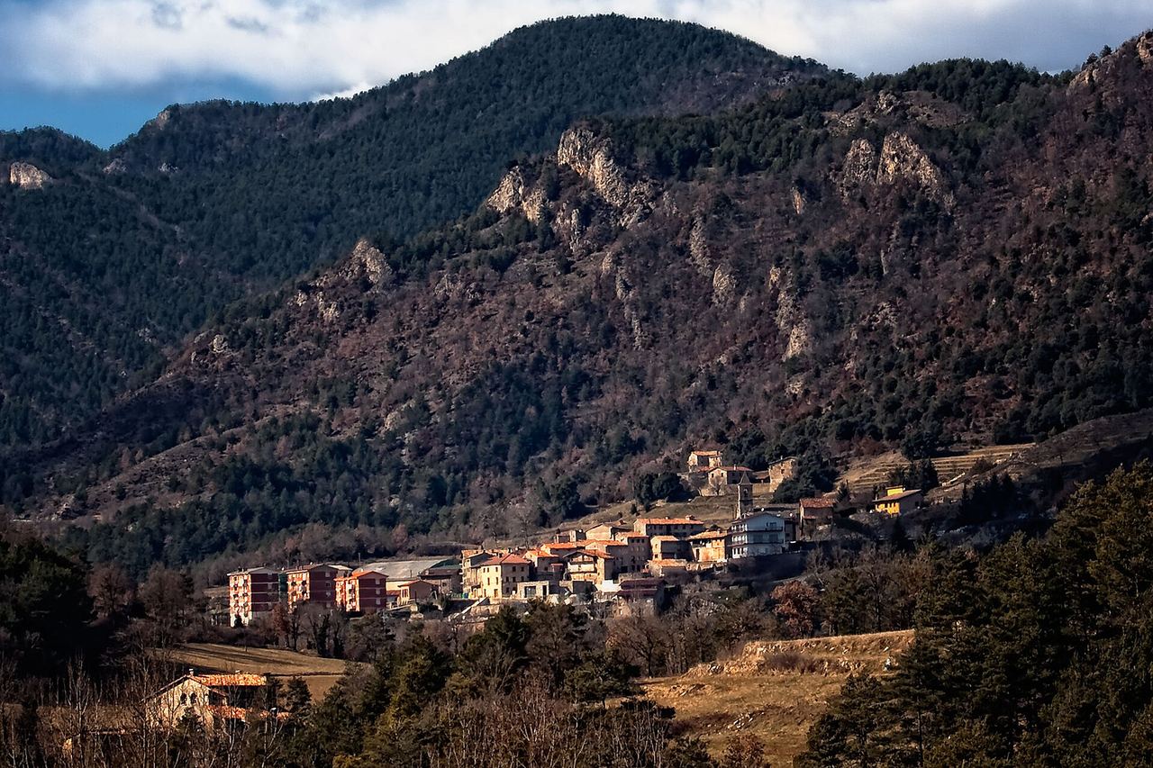 Gombrèn al peu d'una muntanya boscosa amb cingles de roca: cases de teulada vermella i, al davant, prats secs i arbres