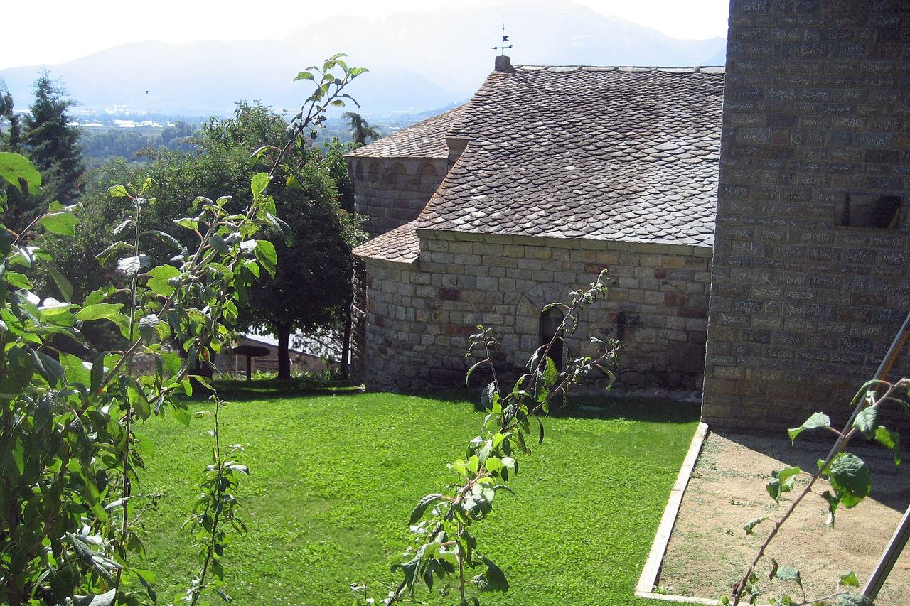 Església de pedra de Sant Miquel d'Isòvol, amb absis i teulada de lloses, gespa al davant i muntanyes entre la boirina