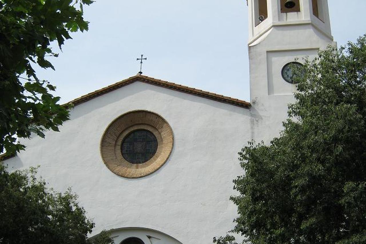 Façana blanca de l'església de Sant Esteve de la Selva de Mar, amb el rosetó i el campanar amb rellotge, entre arbres