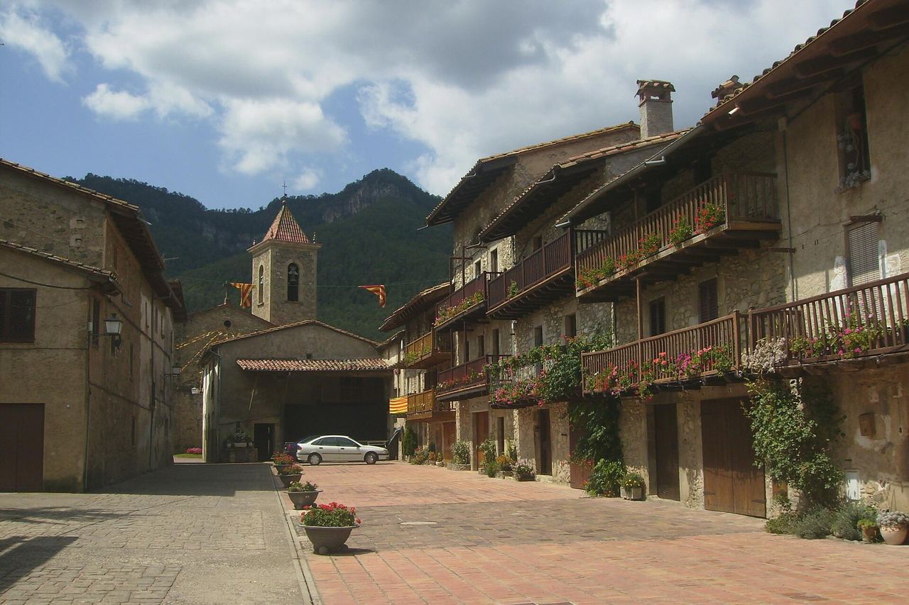 El carrer Teixeda dels Hostalets d'en Bas, a la Vall d'en Bas, amb balcons de fusta plens de flors i el campanar al fons