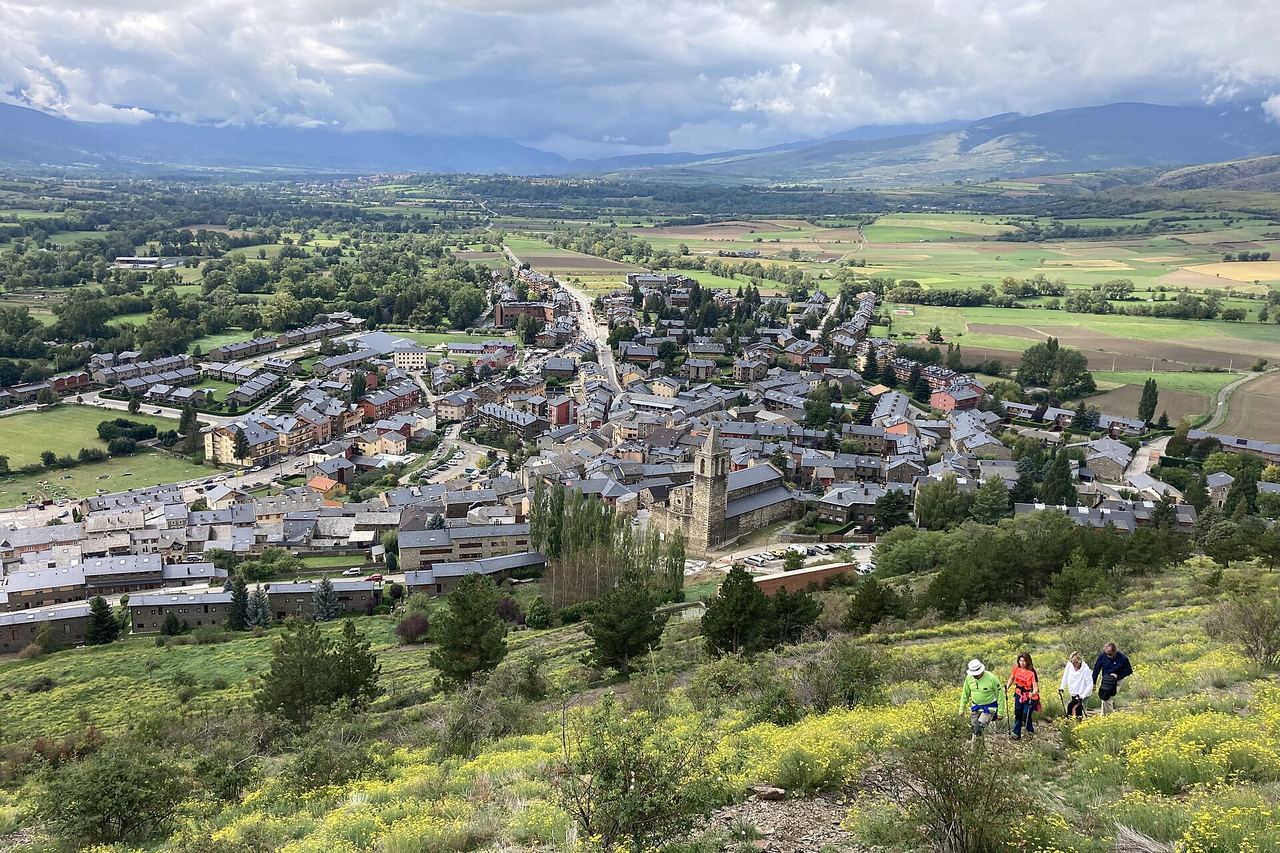 Llívia vista des de dalt: cases i església al mig, la plana amb camps al fons i quatre excursionistes entre flors grogues