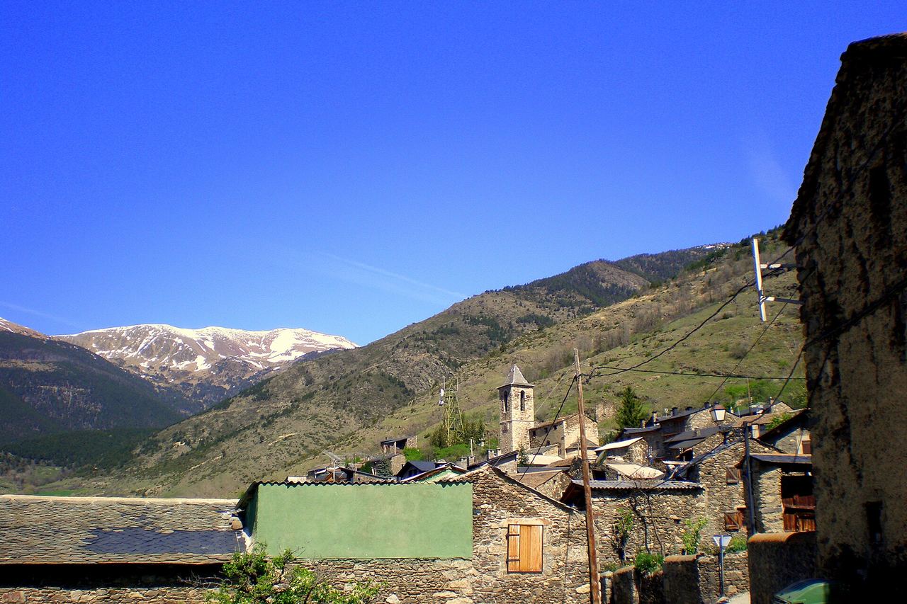 Meranges: el campanar i cases de pedra i pissarra, un mur verd al davant i cims nevats al fons, sota un cel blau