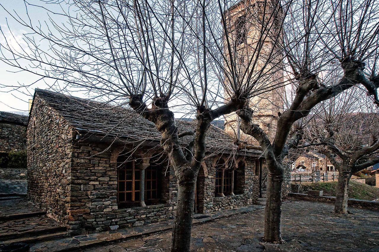 Planoles: edifici de pedra amb teulada de llosa i finestres d'arc, un campanar al darrere i arbres sense fulles