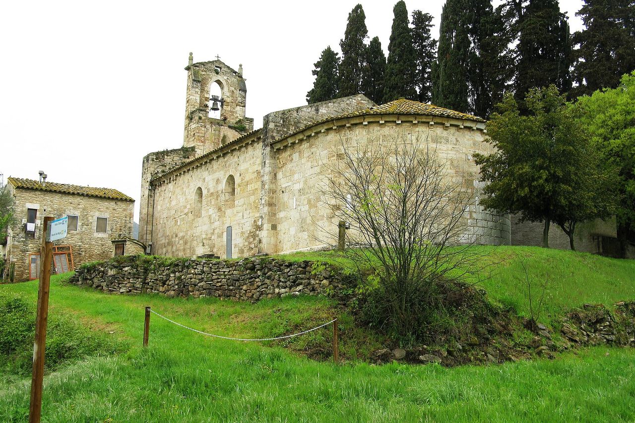 L'església romànica de Santa Maria de Porqueres, amb l'absis i el campanar d'espadanya, entre xiprers i un prat verd