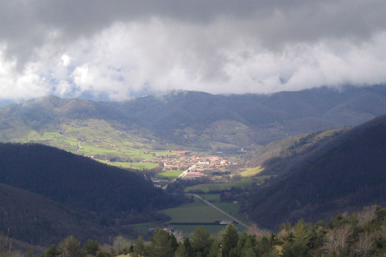El poble de Camprodon, il·luminat pel sol al fons de la vall, entre prats i muntanyes boscoses sota núvols baixos