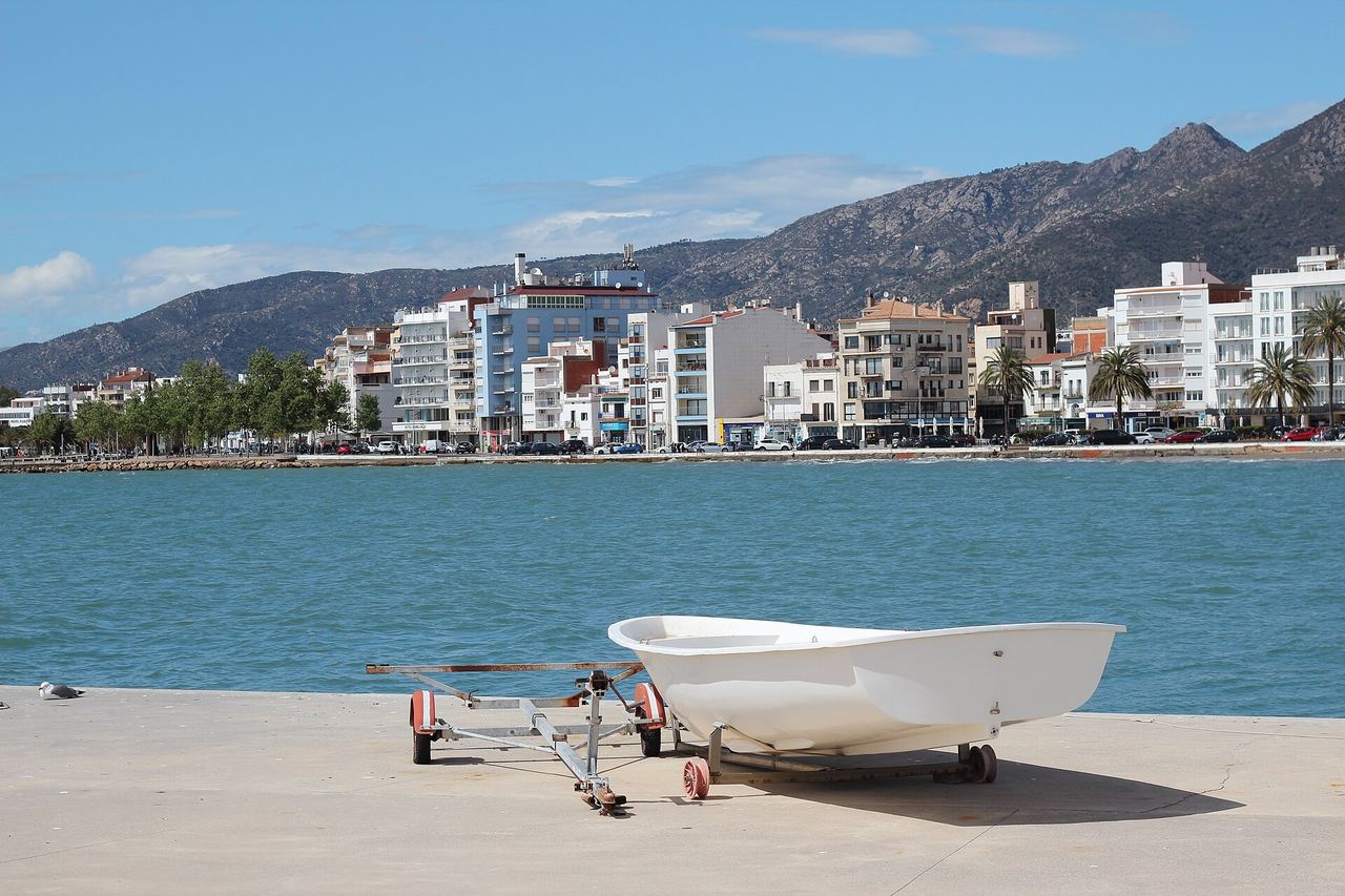 El paseo marítimo de Roses visto desde el muelle, con una barca en primer plano y las montañas al fondo