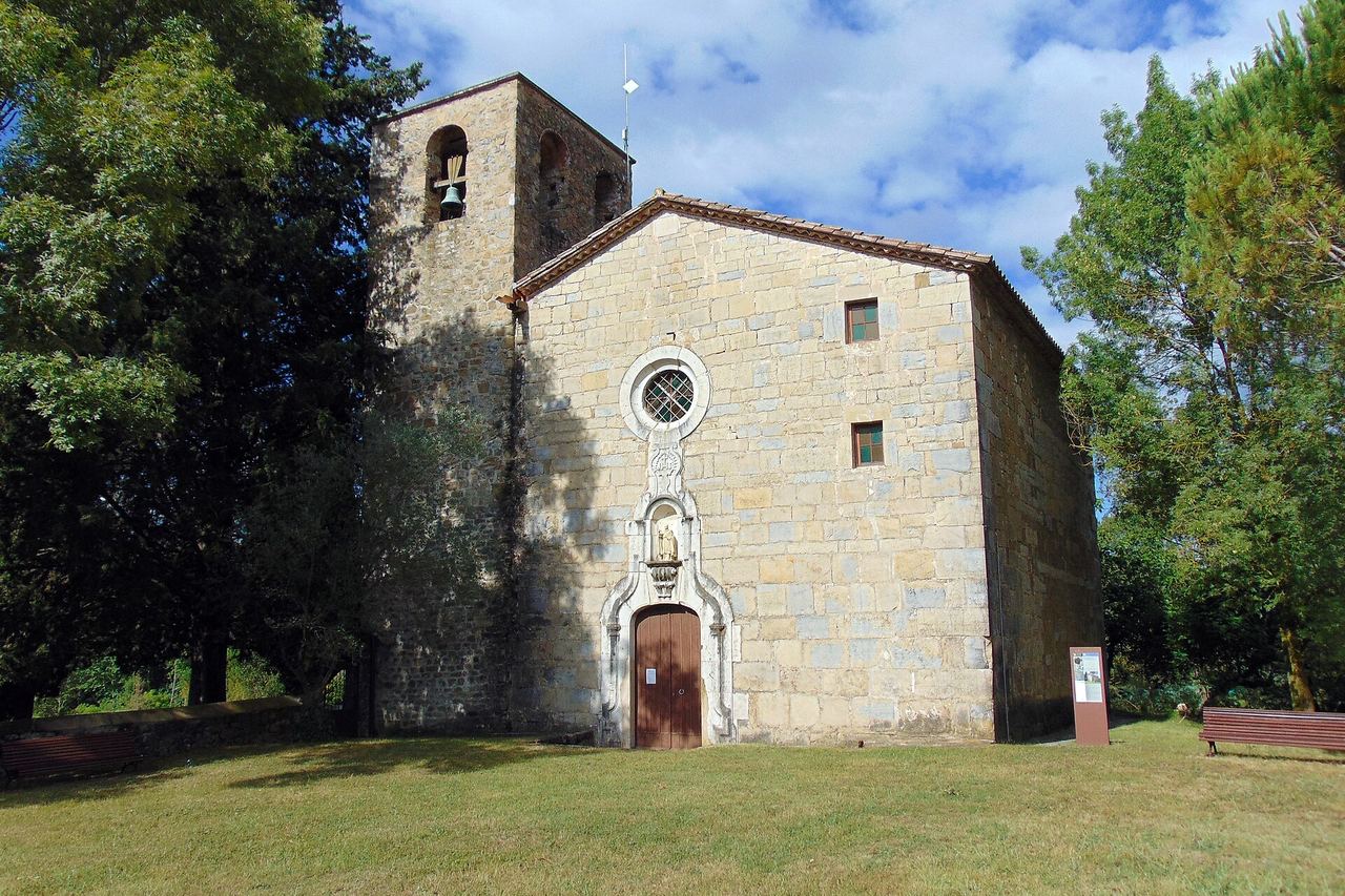 Església de pedra amb campanar, envoltada d'arbres, amb gespa i bancs al davant, al terme de Sales de Llierca