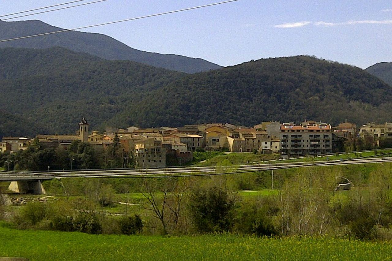 Sant Jaume de Llierca vist des dels camps: el campanar i les cases, una carretera al davant i muntanyes boscoses al darrere