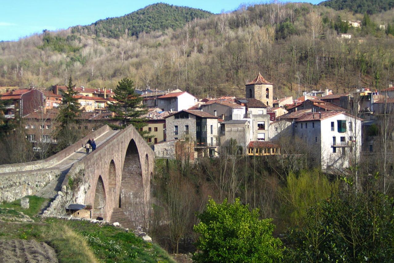 Pont de pedra d'arc apuntat amb dues persones que el travessen i cases de Sant Joan de les Abadesses sota un turó de bosc