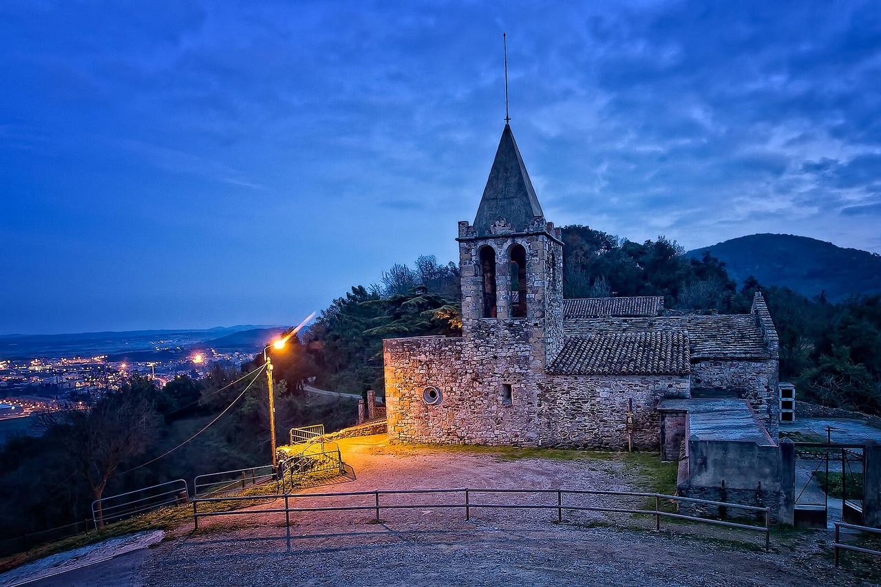 L'església de pedra de Sant Julià de Ramis al vespre, amb el campanar, un fanal encès i una ciutat il·luminada a la plana