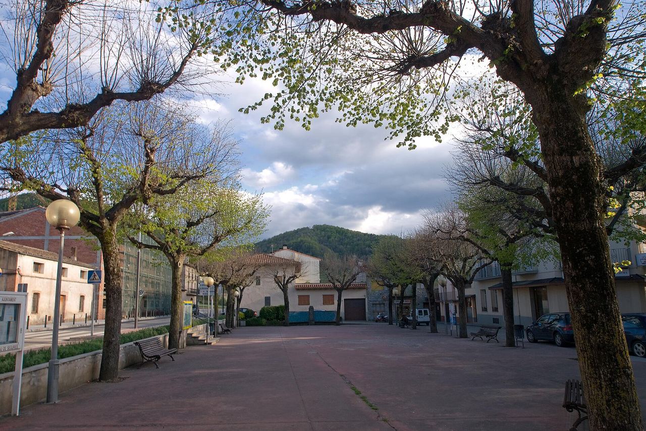 Plaça de Sant Pau de Segúries amb arbres grans, bancs i fanals, cases al voltant i una muntanya de bosc al fons