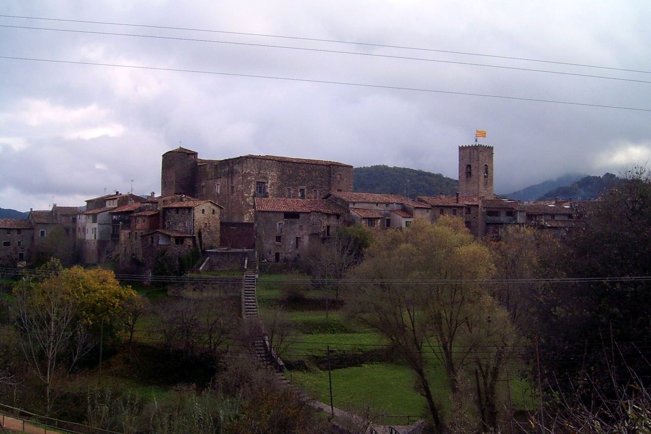 Santa Pau sota un cel ennuvolat: el castell, el campanar amb la senyera i les cases de pedra