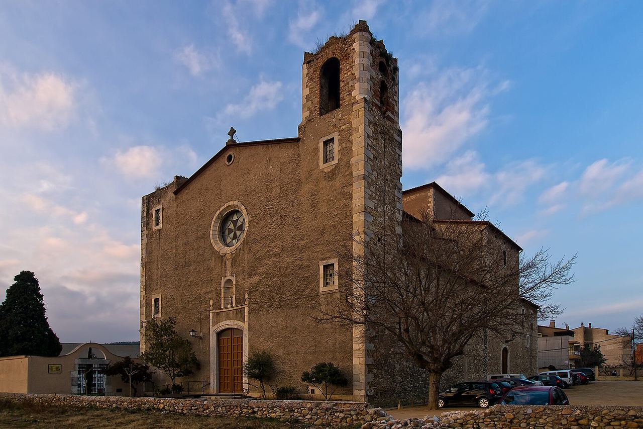 Façana de l'església de Sarrià de Ter, amb rosassa i campanar quadrat, un arbre sense fulles i un mur de pedra al davant