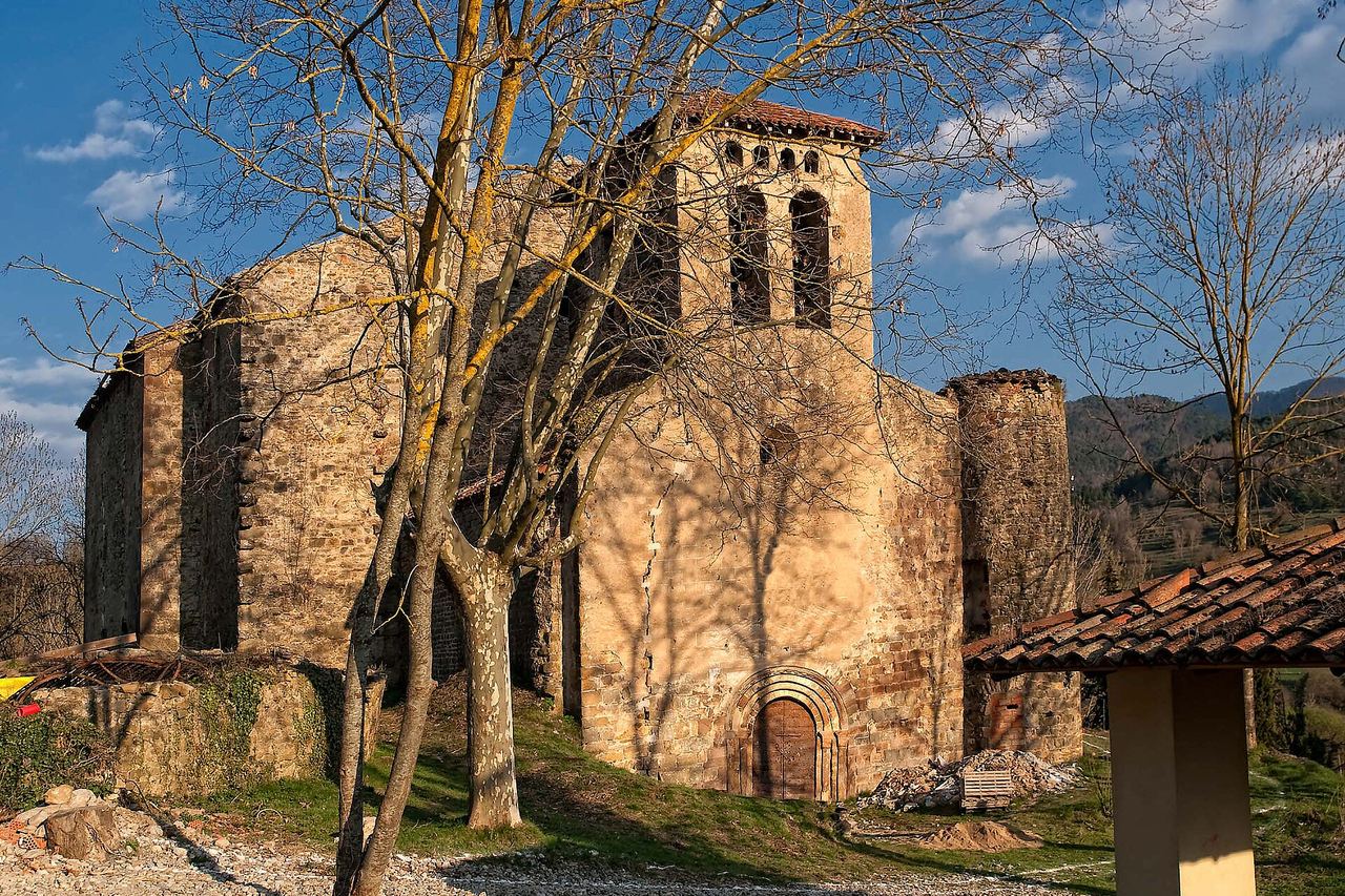 L'església vella de Vallfogona de Ripollès, de pedra, amb campanar, torre rodona i porta d'arc, entre arbres sense fulles