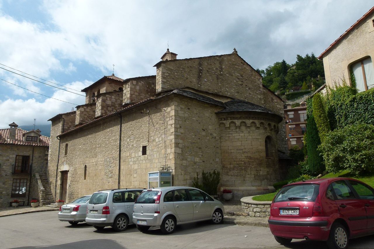 Església de pedra de Vilallonga de Ter amb l'absis arrodonit i cotxes aparcats davant, sota un cel amb núvols